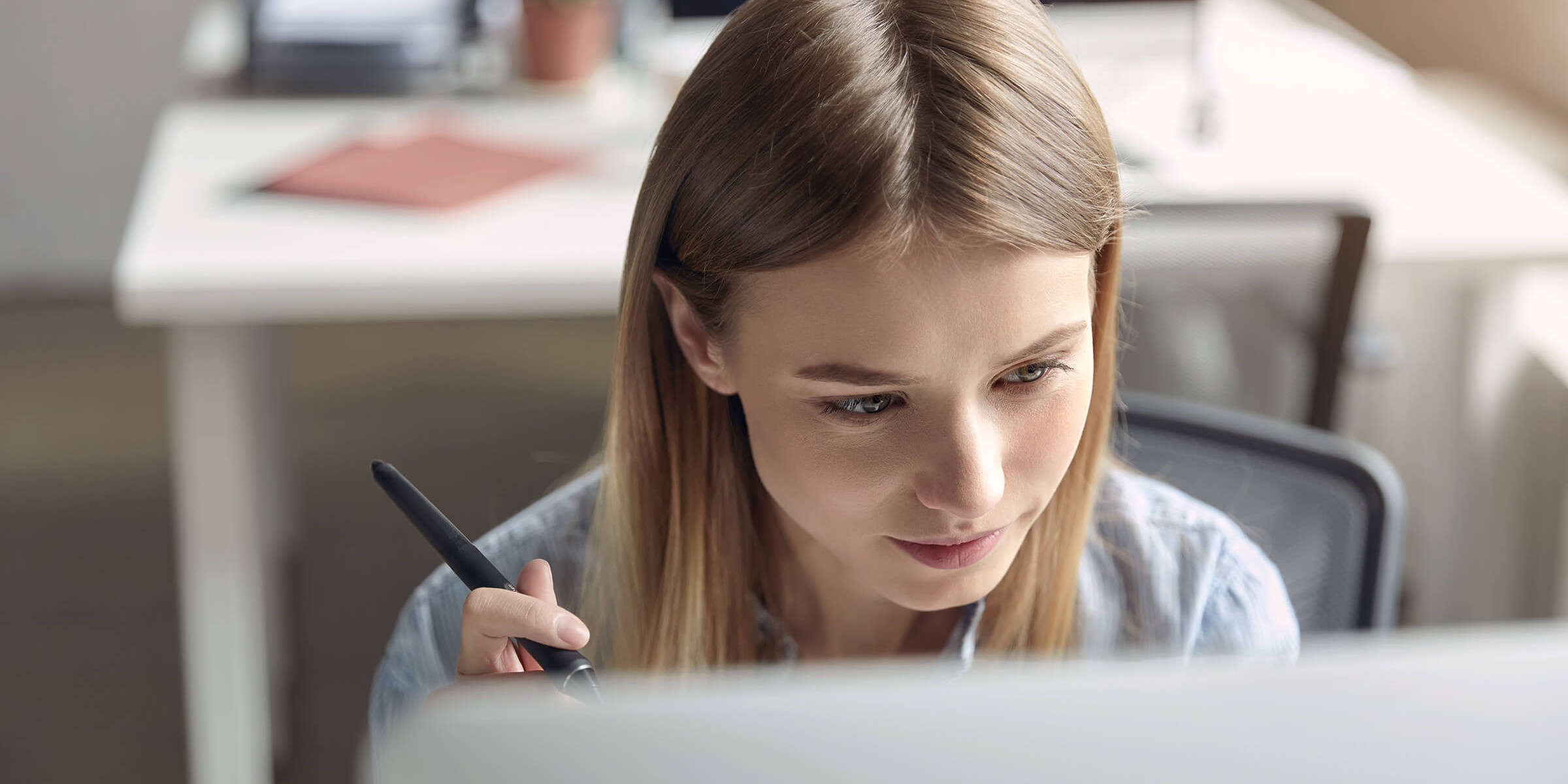 Eine junge Frau sitzt konzentriert an einem Schreibtisch und arbeitet mit einem Grafiktablet. Sie hält einen digitalen Stift in der Hand und schaut aufmerksam auf den Bildschirm. Die Szene vermittelt einen kreativen und produktiven Arbeitsprozess, wie ihn Studierende an der Brand University erleben, um ihre gestalterischen Fähigkeiten weiterzuentwickeln.