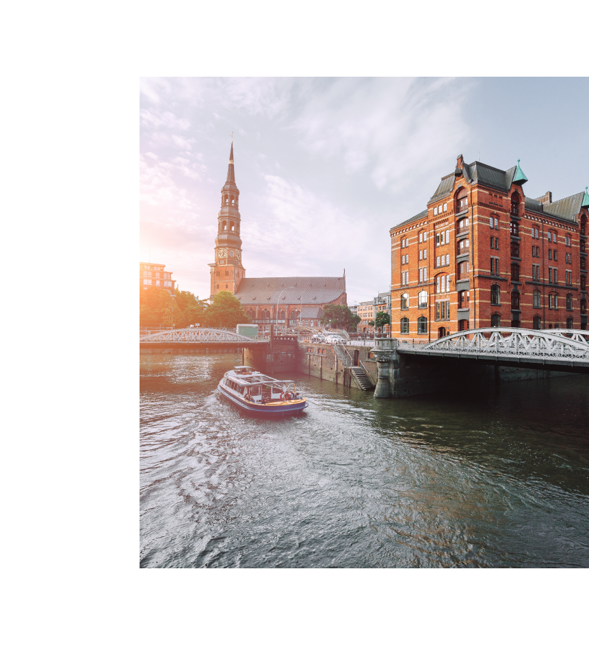 Eine idyllische Szene in der Speicherstadt von Hamburg mit Blick auf einen historischen Backsteinbau und eine Brücke über einen Kanal. Im Vordergrund fährt ein Boot durch das Wasser, und im Hintergrund ist die Kirche St. Katharinen mit ihrem markanten Turm zu sehen. Diese Umgebung, die für ihre architektonische Schönheit bekannt ist, bildet den inspirierenden Standort der Brand University of Applied Sciences.